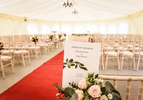 Elegant wedding marquee setup with floral decor and ceremony programme on a chair.