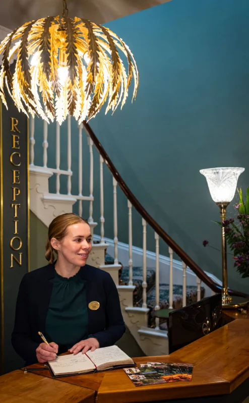 Warm reception at Fernhill House Hotel with staff member, staircase, and elegant lighting.