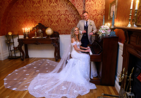 Couple in wedding attire at piano in elegant, warmly lit room with vintage décor.