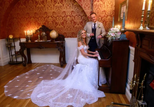 Couple in wedding attire by piano in elegant, vintage-style room at Fernhill House Hotel.