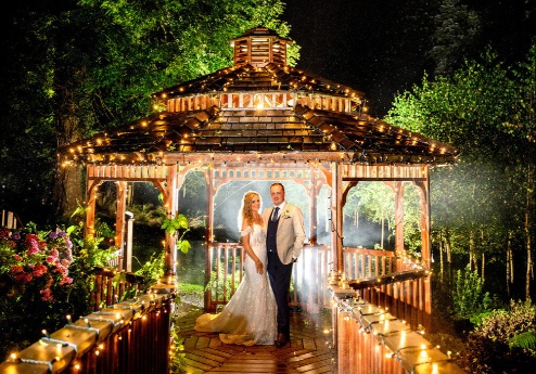 A couple stands on a lit-up wooden bridge in front of a garden gazebo surrounded by trees.