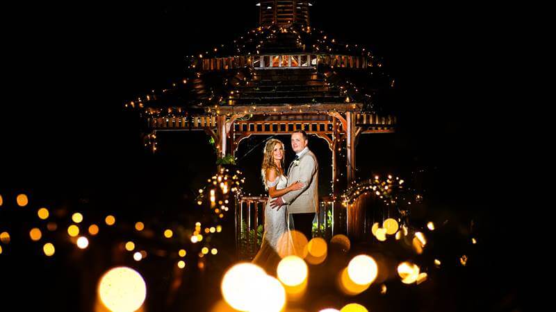 A couple embraces beneath a lit gazebo at night, surrounded by glowing fairy lights.