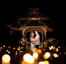 A smiling couple, illuminated by fairy lights, stands under a wooden gazebo at night.