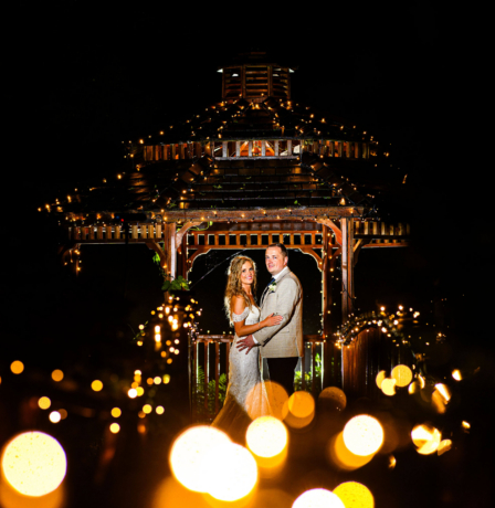 A couple in elegant attire smiling under a gazebo adorned with fairy lights at Fernhill House Hotel.