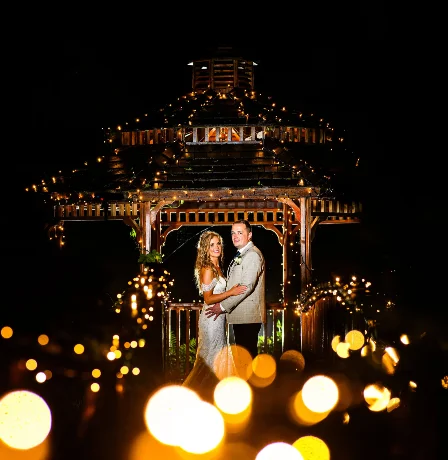 A smiling couple, illuminated by fairy lights, stands under a wooden gazebo at night.