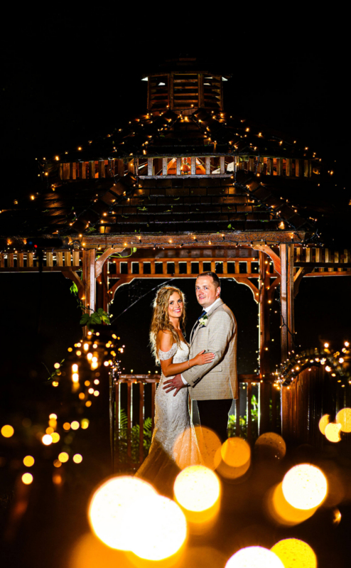 A couple in elegant attire smiling under a gazebo adorned with fairy lights at Fernhill House Hotel.