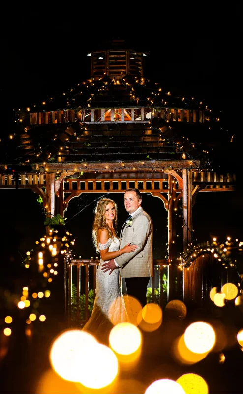 A smiling couple, illuminated by fairy lights, stands under a wooden gazebo at night.