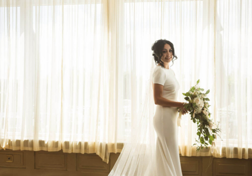 Bride in elegant gown standing by sunlit hotel window