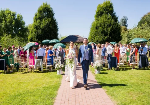 A happy couple walks down an outdoor aisle at Fernhill House Hotel, surrounded by family and friends under clear blue skies.