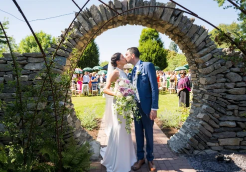 A couple kisses under a stone arch at an outdoor garden wedding with guests looking on.