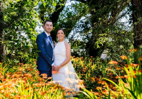 A couple in wedding attire stand in a lush garden with vibrant orange flowers.
