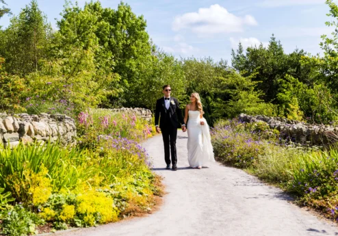 A couple walks hand-in-hand along a flower-lined path in a lush garden.