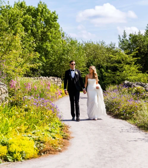 A couple walks hand-in-hand along a flower-lined path in a lush garden.