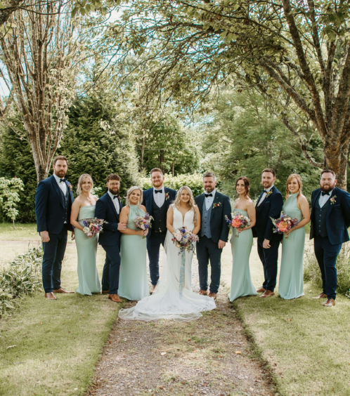 A wedding party smiling under trees in Fernhill House Hotel's lush garden.