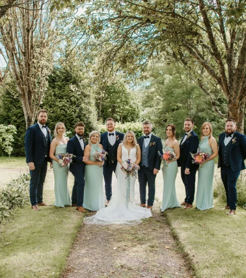 A wedding group stands under trees in Fernhill House Hotel’s lush garden.