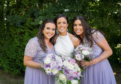 Bride with bridesmaids in lavender dresses joyfully holding bouquets in a lush garden.
