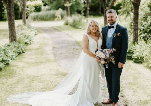 Couple in wedding attire smiling, standing on a garden path surrounded by lush greenery.