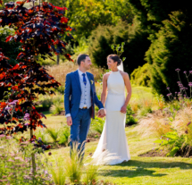 A couple in elegant attire walking hand-in-hand through lush gardens at Fernhill House Hotel.