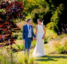 A couple in wedding attire strolls through Fernhill House Hotel's lush garden, surrounded by vibrant greenery and colourful flowers.