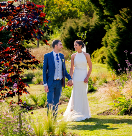A couple in elegant attire walking hand-in-hand through lush gardens at Fernhill House Hotel.