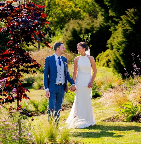 A couple in wedding attire strolls through Fernhill House Hotel's lush garden, surrounded by vibrant greenery and colourful flowers.