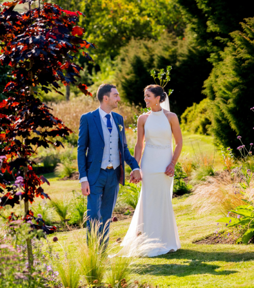 A couple in elegant attire walking hand-in-hand through lush gardens at Fernhill House Hotel.