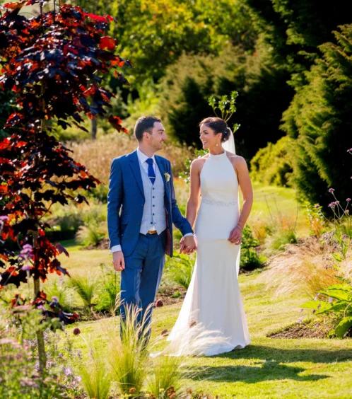 A couple in wedding attire strolls through Fernhill House Hotel's lush garden, surrounded by vibrant greenery and colourful flowers.