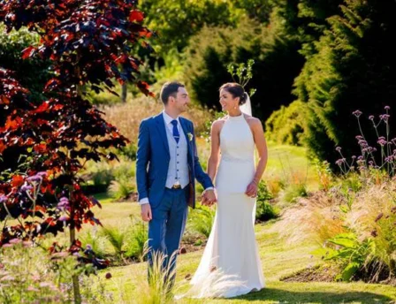 A couple holds hands, strolling through lush gardens at Fernhill House Hotel on a sunny day.
