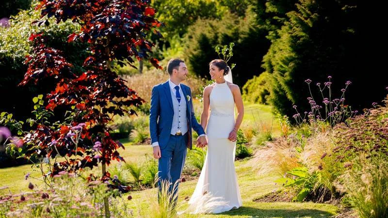 A couple holds hands, strolling through lush gardens at Fernhill House Hotel on a sunny day.