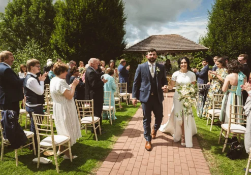 A couple walks down an outdoor aisle after a ceremony, surrounded by applauding family and friends.