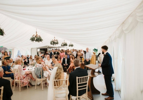 Elegant wedding reception in a bright, decorated marquee with guests listening to a speech.
