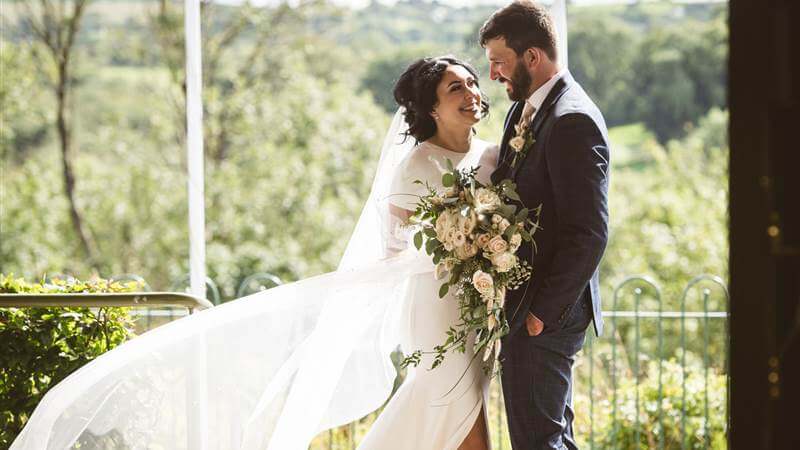 A happy couple in wedding attire embrace outside Fernhill House Hotel, surrounded by lush greenery.