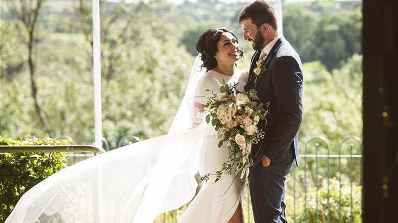 A joyful couple in wedding attire share a moment on a lush garden terrace at Fernhill House Hotel.