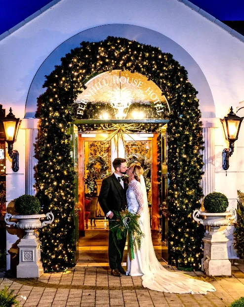 A couple embracing under festive lights at Fernhill House Hotel entrance at night.