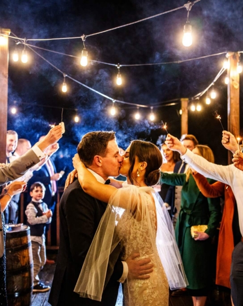 A couple kisses under string lights, surrounded by family holding sparklers at Fernhill House Hotel.
