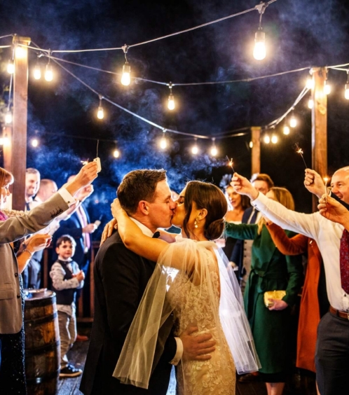 A couple kisses under string lights, surrounded by family holding sparklers at Fernhill House Hotel.