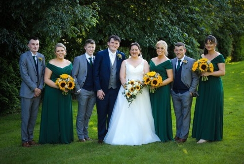 A joyful wedding party poses with sunflowers in a lush garden setting.