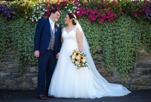 A couple on their wedding day, smiling in front of a floral wall at Fernhill House Hotel.