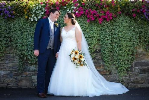 Couple in wedding attire smiling by a floral wall at Fernhill House Hotel.