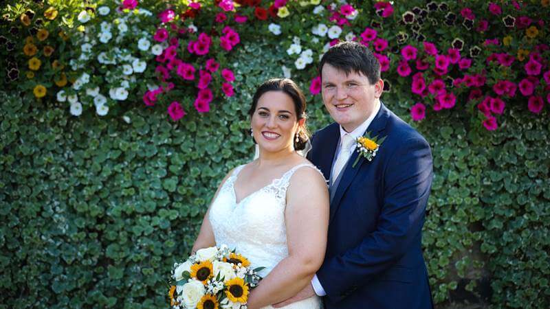 Couple smiling at Fernhill House Hotel garden in front of vibrant flower wall.