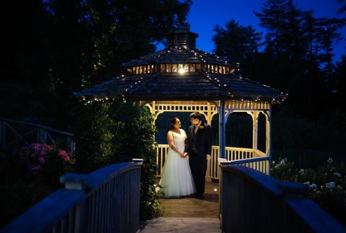 A couple stands under a lit gazebo in a garden at dusk, surrounded by lush greenery.