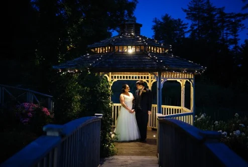 A couple standing under a lit gazebo at night in a scenic garden setting.