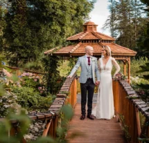 Couple holding hands on a charming wooden bridge, with a gazebo and lush garden backdrop.