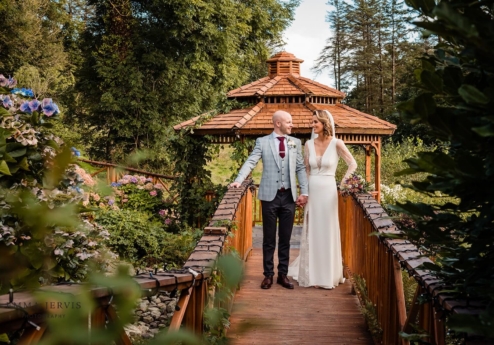 A couple holds hands on a bridge in lush gardens at Fernhill House Hotel, with a gazebo behind them.