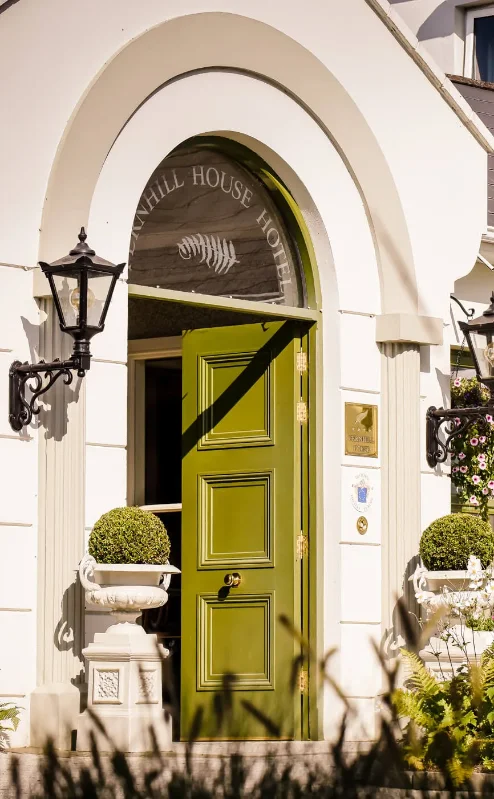 Elegant entrance of Fernhill House Hotel on a sunny day, with lush greenery and welcoming atmosphere.