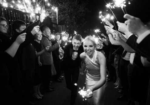A joyful couple holds sparklers, surrounded by smiling guests, celebrating in evening light.