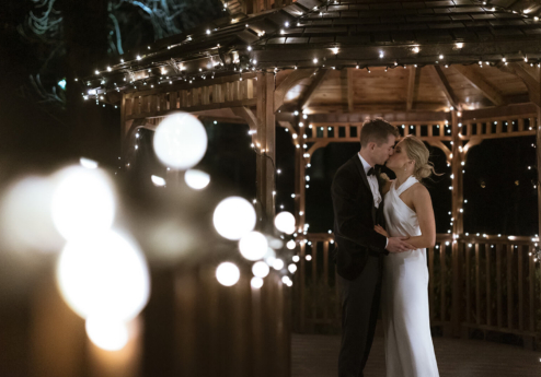 A couple sharing a kiss in a warmly lit gazebo decorated with fairy lights at night.