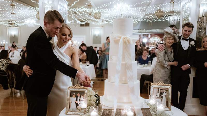 A joyful couple cutting a wedding cake in a warmly lit reception hall, surrounded by family.