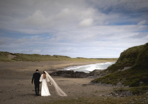 A couple walks hand in hand on a tranquil beach with grassy dunes and gentle waves.