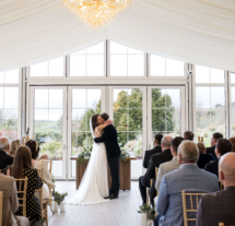 A couple celebrates their wedding with a kiss in a bright, elegant conservatory, surrounded by family and friends.